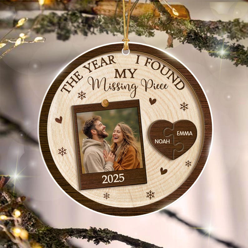 A round wooden ornament featuring the text THE YEAR I FOUND MY Missing Piece and 2025. It displays a square photo of a couple, a heart-shaped puzzle piece graphic with the names Noah and Emma, and small snowflake and heart icons.