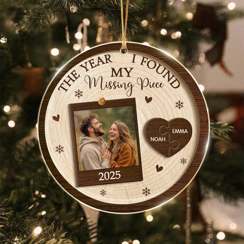 A round wooden Christmas ornament featuring the text THE YEAR I FOUND MY Missing Piece. It includes a central photo frame with a couple and the year 2025, plus a heart-shaped puzzle piece ornament with the names Noah and Emma.