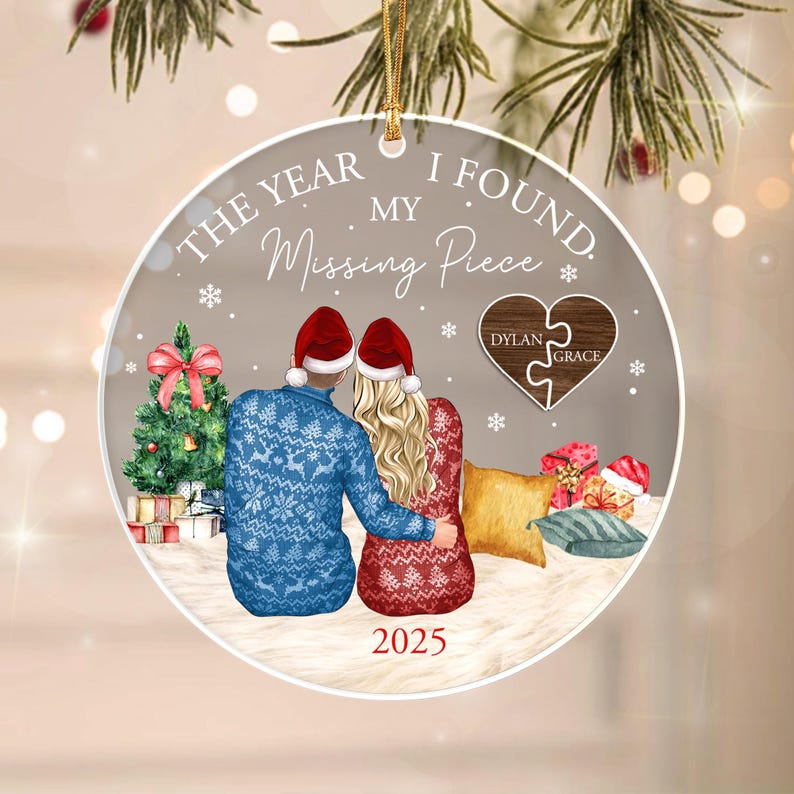 A round Christmas ornament featuring a couple in red hats and patterned sweaters sitting together. The text reads THE YEAR I FOUND MY Missing Piece and 2025. A heart-shaped puzzle piece graphic displays the names Dylan and Grace.