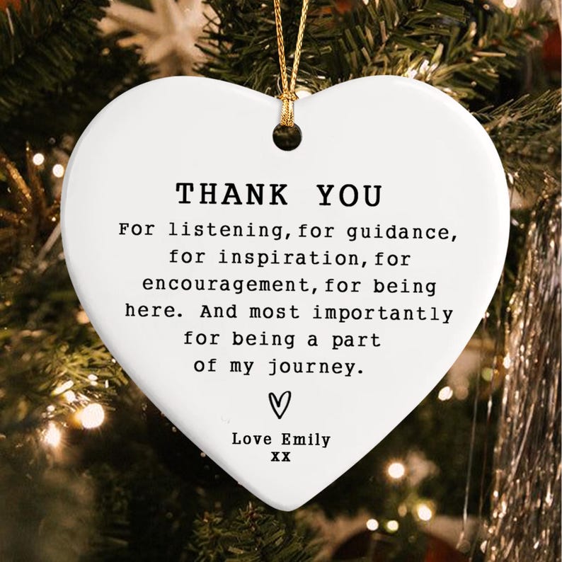 A white heart-shaped ornament with the words "THANK YOU" in black text. The ornament includes a message of gratitude and is suspended by a gold string. The background is a blurred Christmas tree.