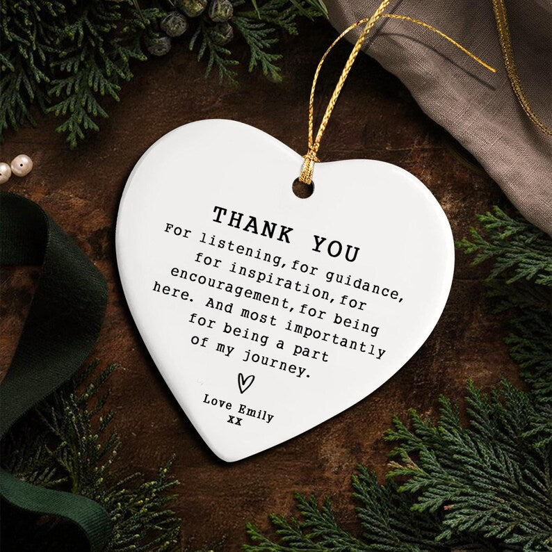 A white heart-shaped ornament with gold string. The ornament has the words "THANK YOU" and a message of gratitude printed in black. The ornament is surrounded by greenery and a dark brown surface.