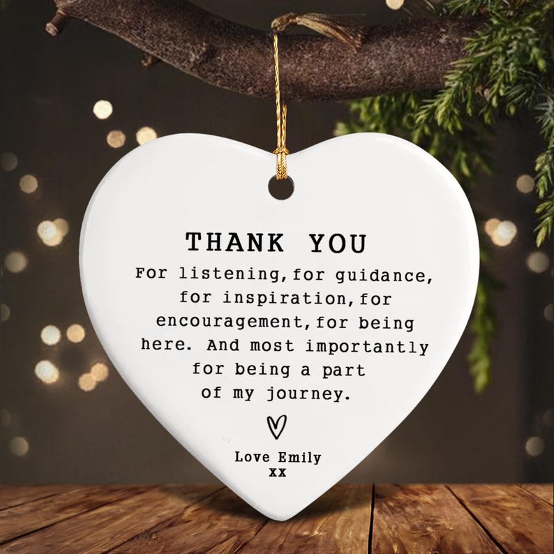 White heart-shaped ceramic ornament with gold hanging string. The ornament has the words "THANK YOU" and a message of gratitude, including "Love Emily" at the bottom. The ornament is hanging from a tree branch.