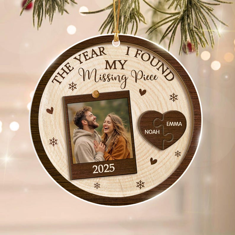 A round wooden Christmas ornament featuring the text THE YEAR I FOUND MY Missing Piece and 2025. It includes a square photo frame with a couple, a heart-shaped puzzle piece with the names Noah and Emma, and small snowflake icons.