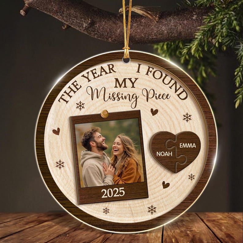 A round wooden ornament featuring a photo of a couple, the text "THE YEAR I FOUND MY Missing Piece" and "2025", and a heart-shaped puzzle piece graphic with the names "NOAH" and "EMMA" printed on it, suspended by a gold cord.