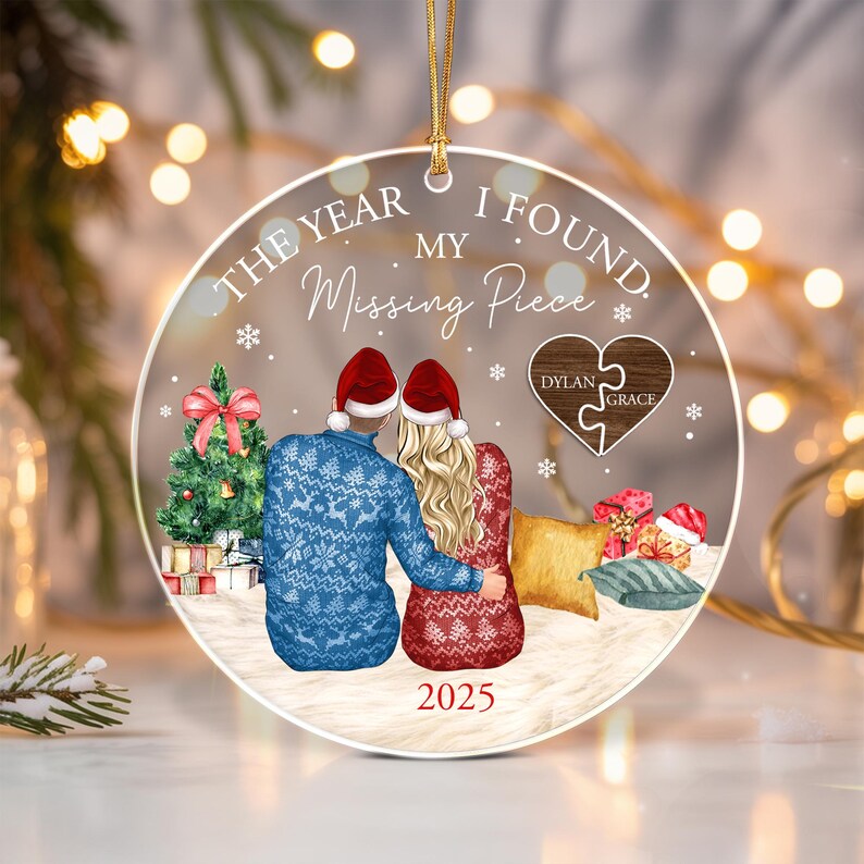 A round clear acrylic Christmas ornament featuring a couple in Santa hats sitting together. The text reads THE YEAR I FOUND MY Missing Piece and 2025. A wooden heart puzzle piece is printed with the names Dylan and Grace.
