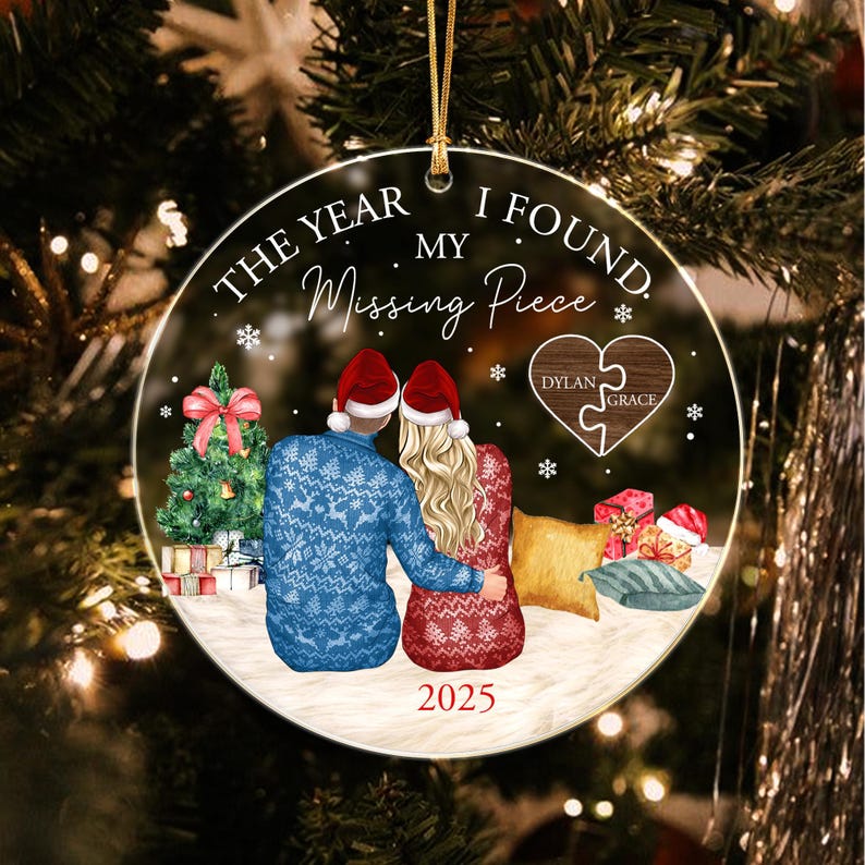 A round Christmas ornament featuring a couple in Santa hats and patterned sweaters sitting together. Text reads THE YEAR I FOUND MY Missing Piece and 2025. A heart-shaped puzzle piece graphic displays the names Dylan and Grace.