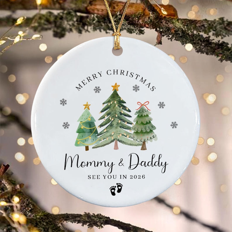 A round white ceramic Christmas ornament featuring three watercolor-style pine trees, gold stars, and snowflakes. The text reads MERRY CHRISTMAS, Mommy & Daddy, SEE YOU IN 2026, with two small black baby footprints at the bottom.