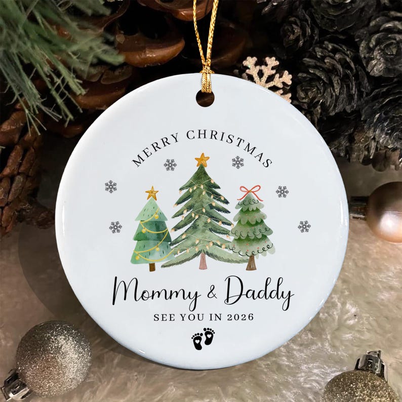 A round white ceramic Christmas ornament featuring three watercolor-style pine trees, the text MERRY CHRISTMAS, Mommy & Daddy, SEE YOU IN 2026, and a small pair of black baby footprints.