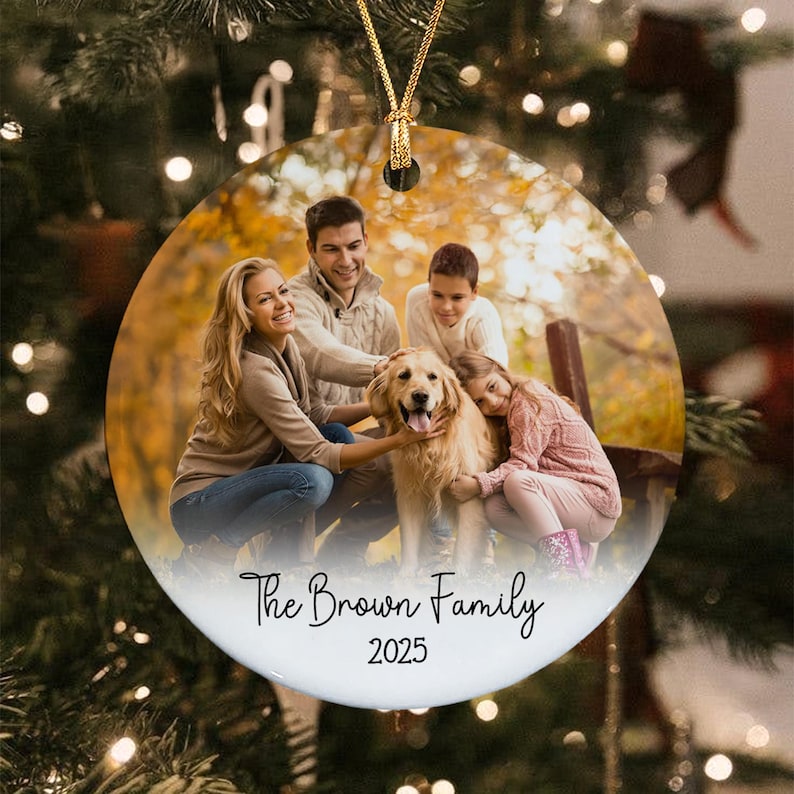 A round ceramic Christmas ornament featuring a printed photograph of a family of four and a golden retriever dog outdoors. The text The Brown Family and the year 2025 are printed in black script and sans-serif font at the bottom.