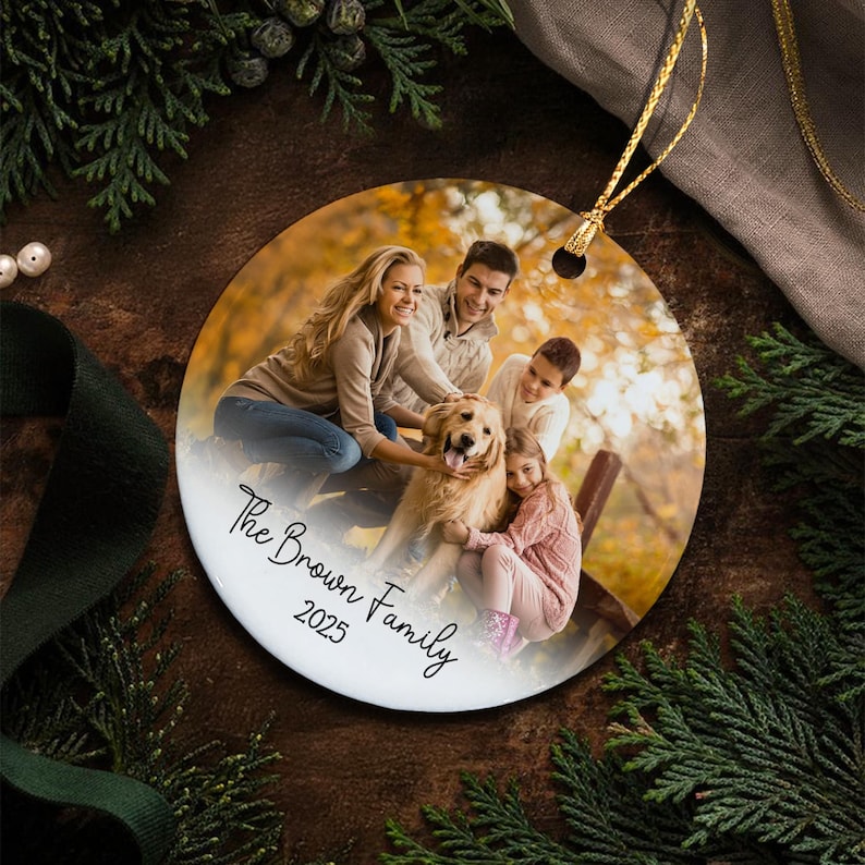 A round ceramic ornament featuring a printed photograph of a family with a golden retriever dog in an autumn setting. The text "The Brown Family 2025" is printed in black script on the lower portion.