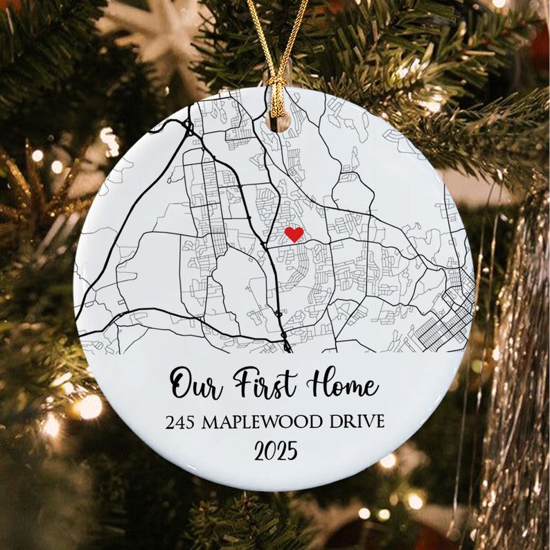 A round white ceramic ornament featuring a black street map graphic with a small red heart icon. The text reads Our First Home, 245 MAPLEWOOD DRIVE, and 2025.