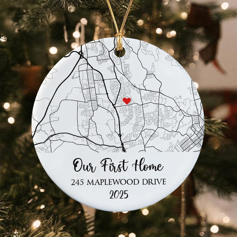 A round white ceramic ornament featuring a black street map print with a small red heart icon. The text reads Our First Home, 245 MAPLEWOOD DRIVE, and 2025. It hangs from a gold string against a blurred green tree background.