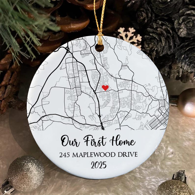 A round white ceramic ornament featuring a black line map with a small red heart icon. The text reads Our First Home, 245 MAPLEWOOD DRIVE, and 2025. It hangs from a gold string and is designed for display as a holiday decoration.