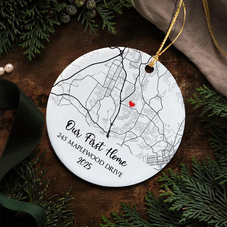 A round white ceramic ornament featuring a black line map print with a small red heart icon. The text reads Our First Home, 245 MAPLEWOOD DRIVE, 2025. It includes a gold hanging string attached through a hole at the top edge.