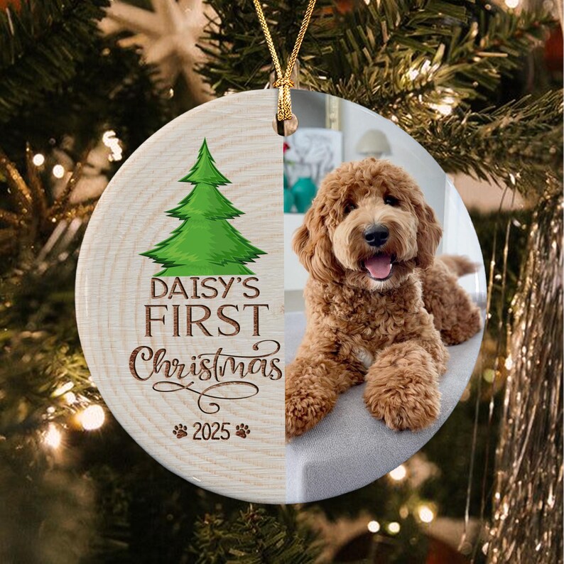A round ceramic Christmas ornament featuring a wood grain pattern on the left side with a green pine tree illustration, the text "DAISY'S FIRST Christmas 2025" with paw prints.