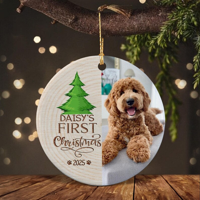 A round ceramic ornament featuring a split design. The left side has a wood grain texture with a green Christmas tree illustration, the text "DAISY'S FIRST Christmas" and "2025" with paw prints.