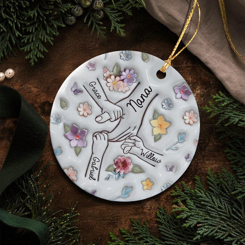 A round white ceramic ornament featuring a central illustration of hands holding together, with the names Grace, Gabriel, and Willow written near each hand and the word Nana in the center, surrounded by colorful floral patterns.