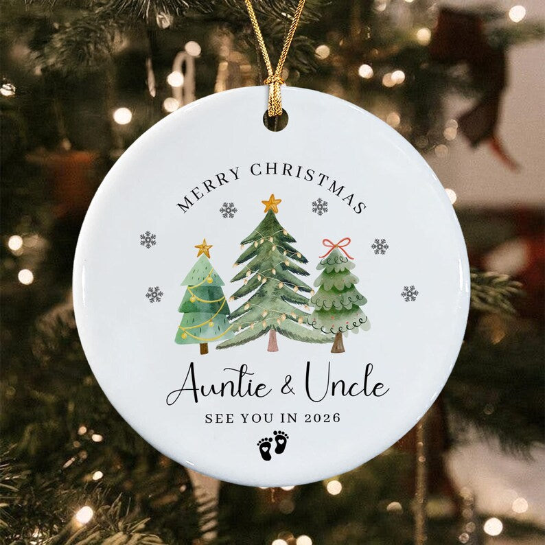 A round white ceramic Christmas ornament featuring a watercolor illustration of three green pine trees with gold stars and a red bow.