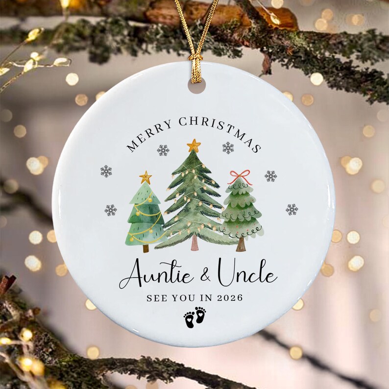 A round white ceramic Christmas ornament featuring three watercolor-style pine trees, gold stars, and snowflakes. The text reads MERRY CHRISTMAS, Auntie & Uncle, SEE YOU IN 2026, with two small black baby footprints at the bottom.