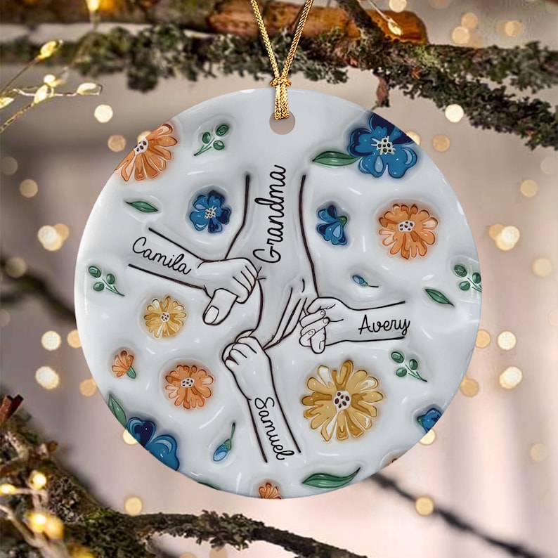 A round white ceramic ornament featuring a central illustration of three hands holding a larger hand. The text "Grandma" is written vertically, with the names "Camila," "Avery," and "Samuel" printed near each hand.