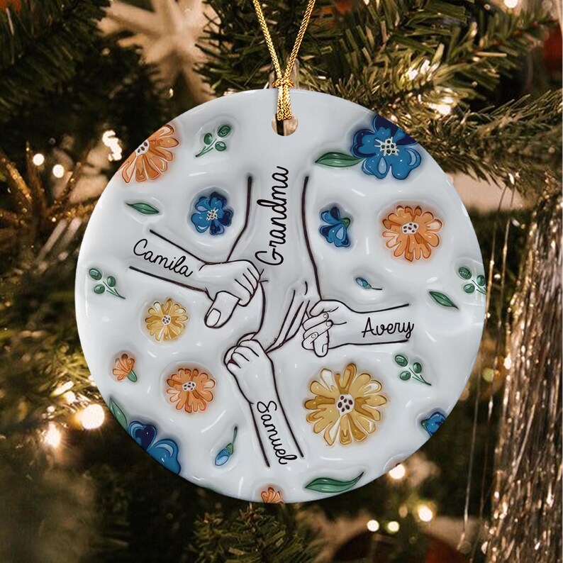 A round white ceramic ornament featuring a central illustration of hands holding together, labeled with the names Camila, Avery, and Samuel, and the word Grandma.