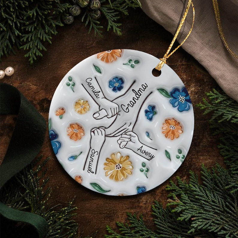 A round white ceramic ornament featuring a central illustration of four hands holding each other. The names Camila, Samuel, and Avery are printed near the hands, with the word Grandma in the center.