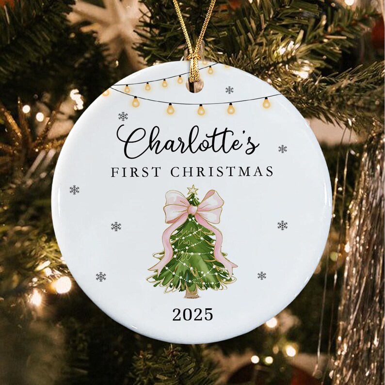 A round white ceramic Christmas ornament featuring a string of lights at the top, the text Charlotte's FIRST CHRISTMAS, a green Christmas tree illustration with a large pink bow, and the year 2025, suspended by a gold cord.