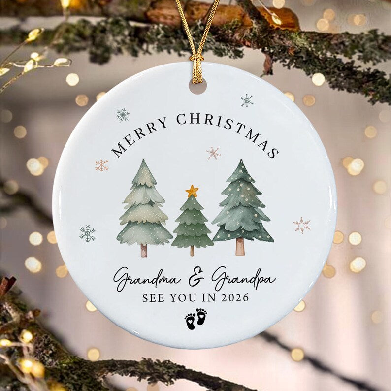 A round white ceramic ornament featuring three watercolor-style pine trees, a gold star, and small snowflakes. The text reads MERRY CHRISTMAS, Grandma & Grandpa, SEE YOU IN 2026, with two small black baby footprints at the bottom.