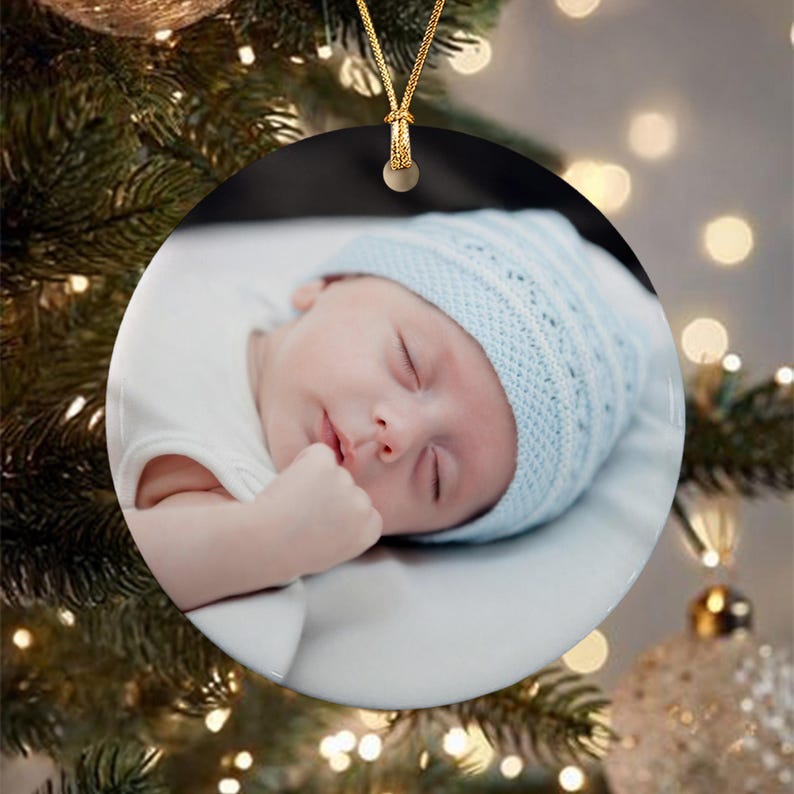 A round ceramic ornament featuring a high-resolution photograph of a sleeping infant wearing a light blue knit cap and a white shirt.