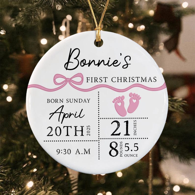 A round white ceramic ornament featuring a pink ribbon graphic and the text Bonnie's FIRST CHRISTMAS. It displays birth details: Sunday, April 20th 2025, 9:30 A.M., 21 inches, and 8 pounds 5.5 ounces, with pink baby footprints.