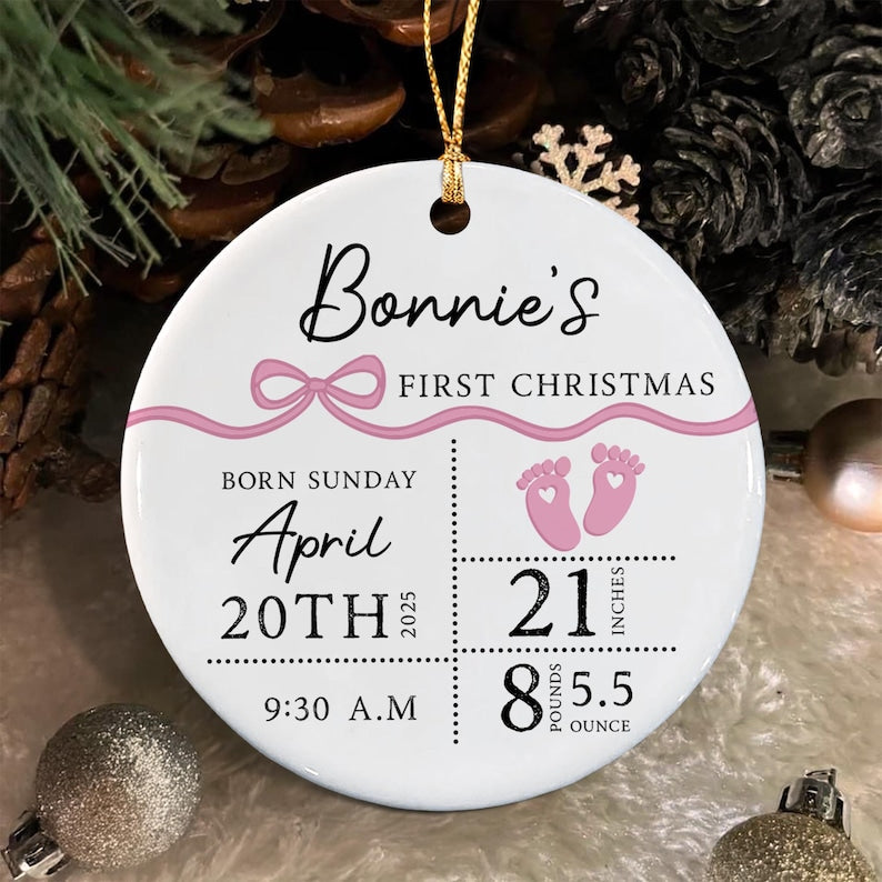 A round white ceramic ornament featuring a pink ribbon graphic and the text Bonnie's FIRST CHRISTMAS. It includes birth details: Sunday April 20th 2025, 9:30 A.M, 21 inches, and 8 pounds 5.5 ounces, with pink baby footprint icons.