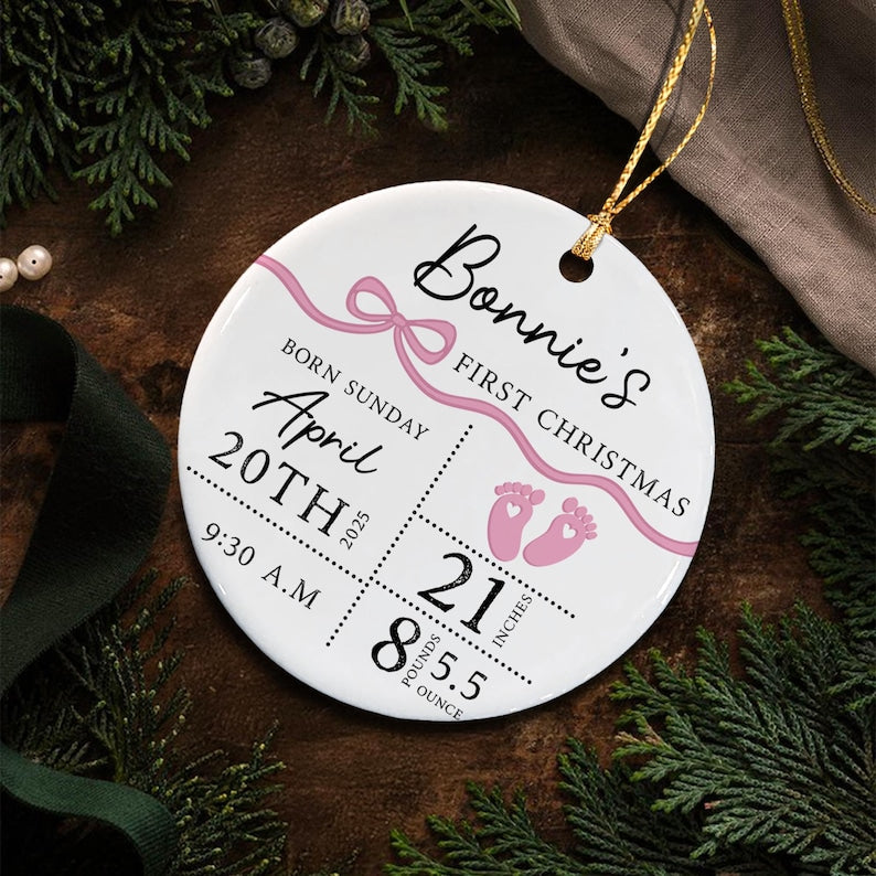 A round white ceramic ornament with a gold hanging string. It features the text Bonnie's First Christmas, a pink ribbon graphic, and two pink baby footprints.