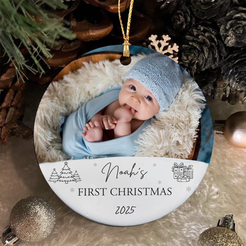 A round ceramic Christmas ornament featuring a photograph of a baby in a light blue knit hat and blanket. The white lower section displays the text Noah's FIRST CHRISTMAS 2025, accompanied by line art of pine trees and gift boxes.
