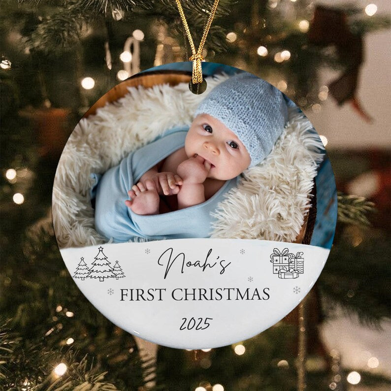 Round Christmas ornament featuring a baby in a blue hat and swaddle. The ornament has the text "Noah's FIRST CHRISTMAS 2025" with Christmas tree and gift illustrations. A gold string is attached for hanging.