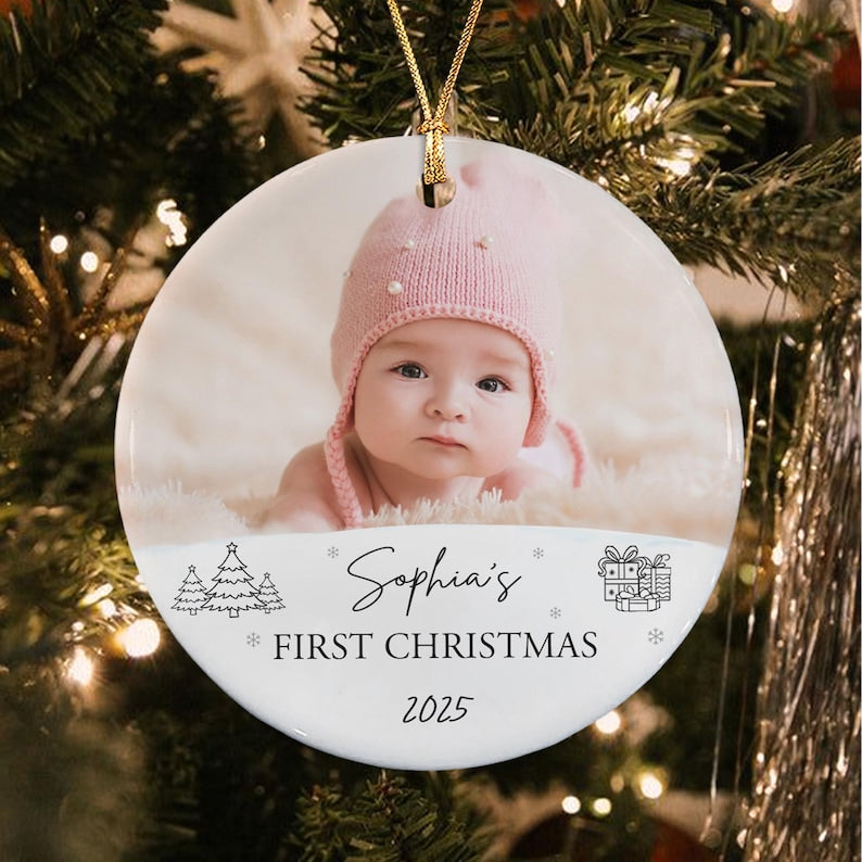 A round white ceramic ornament featuring a photograph of a baby wearing a pink knit hat. The text reads Sophia's FIRST CHRISTMAS 2025, accompanied by black line art illustrations of three pine trees and a stack of gift boxes.