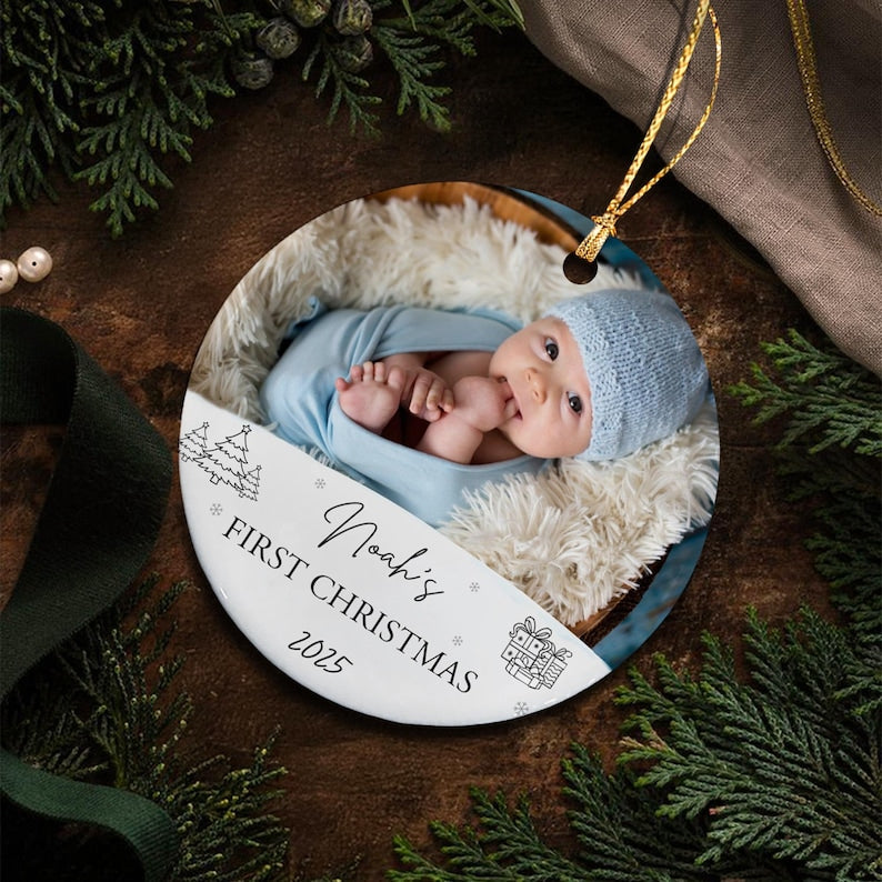 Round Christmas ornament featuring a baby in a blue hat and swaddling. The ornament has the text "Noah's First Christmas 2025" with snowflake and gift box graphics. A gold string is attached for hanging.