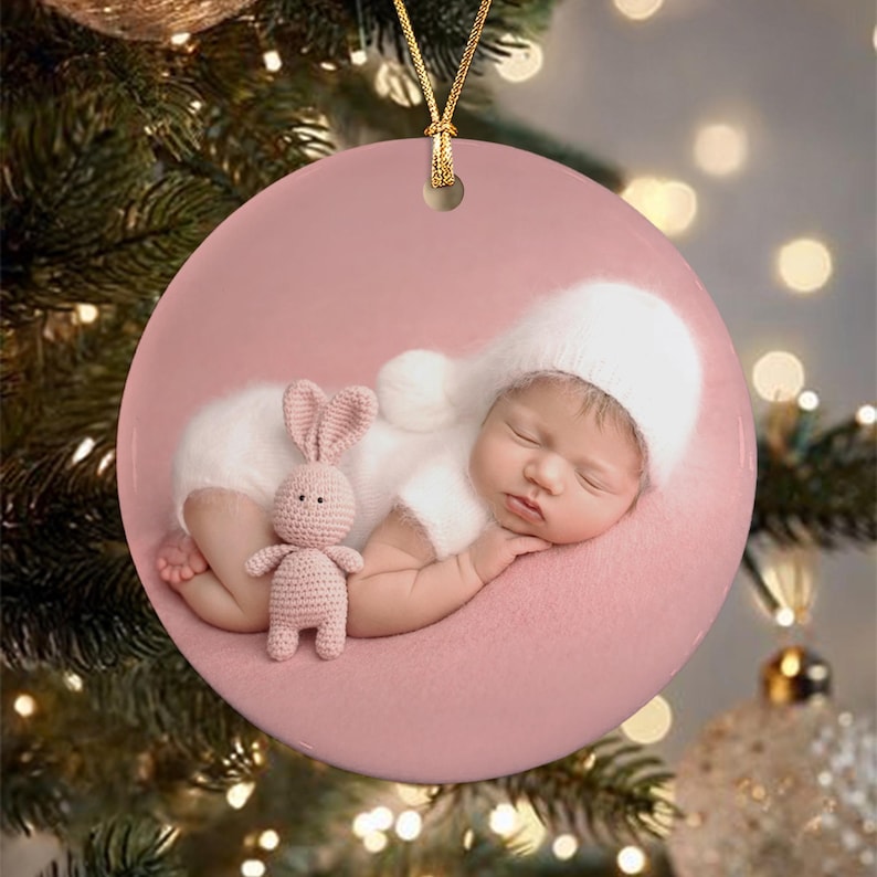 A round ceramic ornament featuring a photograph of a sleeping infant wearing a white knit hat and matching outfit, lying on a soft pink surface while holding a small, light pink crocheted rabbit toy.
