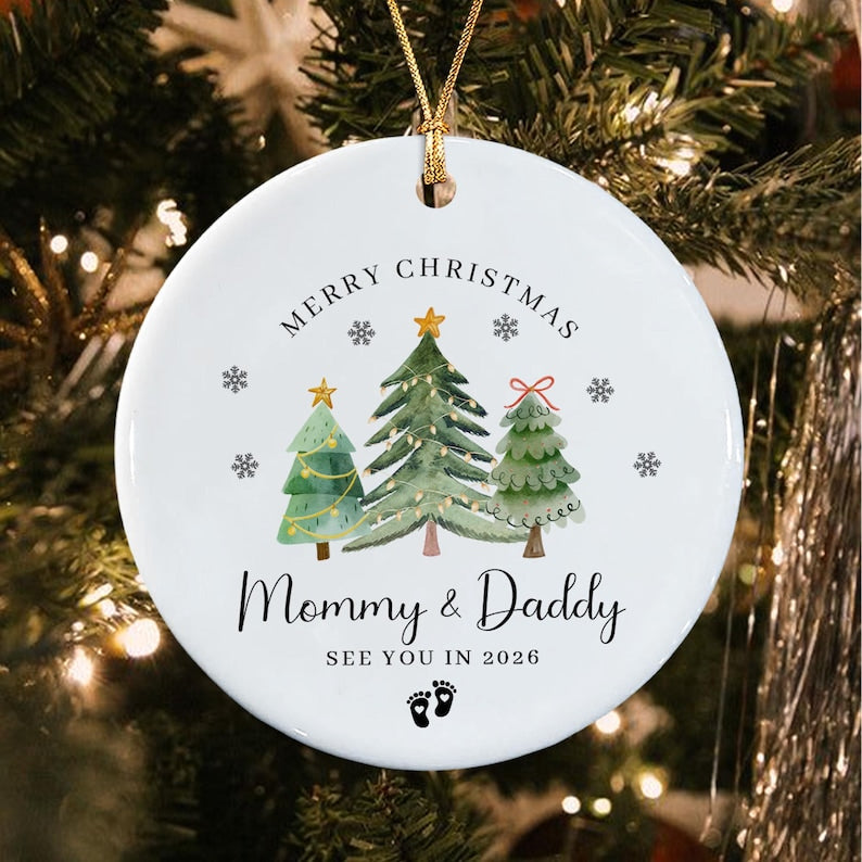 A round white ceramic ornament with a gold string featuring a watercolor illustration of three Christmas trees, snowflakes, and the text Merry Christmas, Mommy & Daddy, SEE YOU IN 2026, and a pair of small black baby footprints.