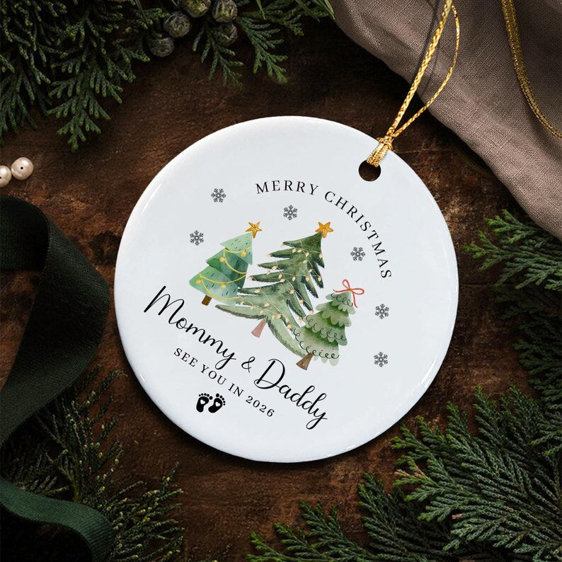 A round white ceramic ornament featuring three watercolor-style green Christmas trees with gold stars. The text reads MERRY CHRISTMAS, Mommy & Daddy, and SEE YOU IN 2026, accompanied by small black baby footprints and snowflakes.
