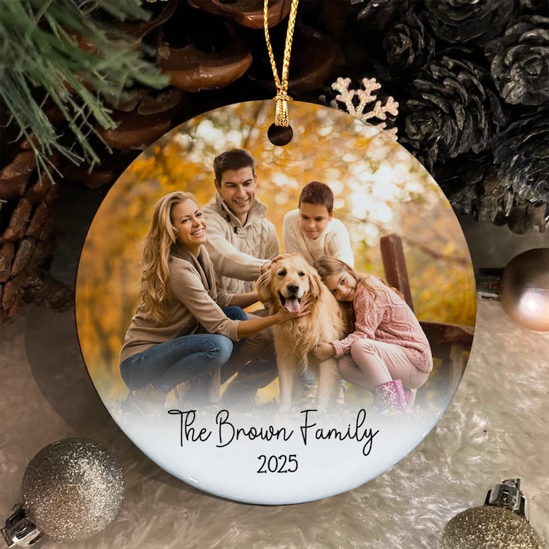A round ceramic Christmas ornament featuring a printed photograph of a family with a golden retriever dog in an autumn setting. The text "The Brown Family" and "2025" is printed in black script and sans-serif font at the bottom.