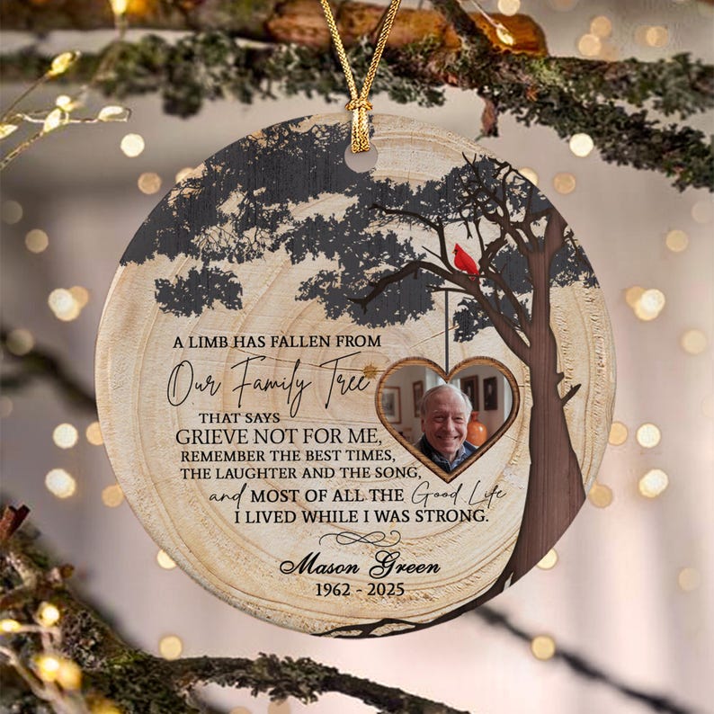 A round ceramic memorial ornament featuring a wood grain pattern, a silhouette of a tree with a red cardinal, and a heart-shaped photo frame. It includes the text A LIMB HAS FALLEN FROM Our Family Tree, Mason Green, 1962-2025.
