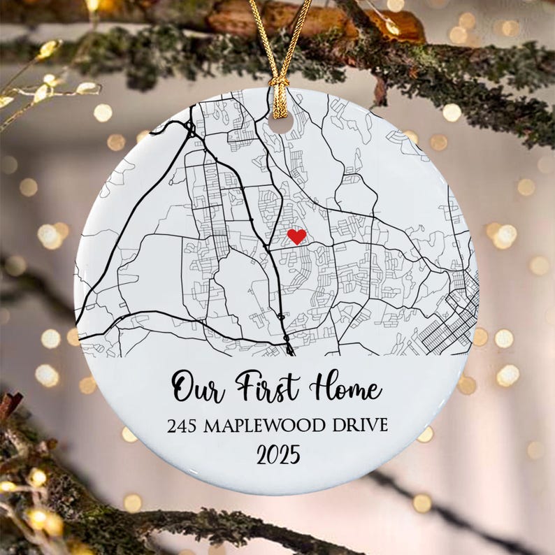 A round white ceramic ornament featuring a black line street map with a small red heart icon. The text reads Our First Home, 245 MAPLEWOOD DRIVE, and 2025. It hangs from a gold string against a blurred background of tree branches.
