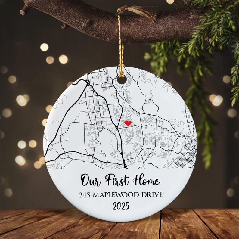 A round white ceramic ornament featuring a black line map graphic with a small red heart icon. The text reads Our First Home, 245 MAPLEWOOD DRIVE, and 2025. It hangs from a gold string against a blurred dark background.