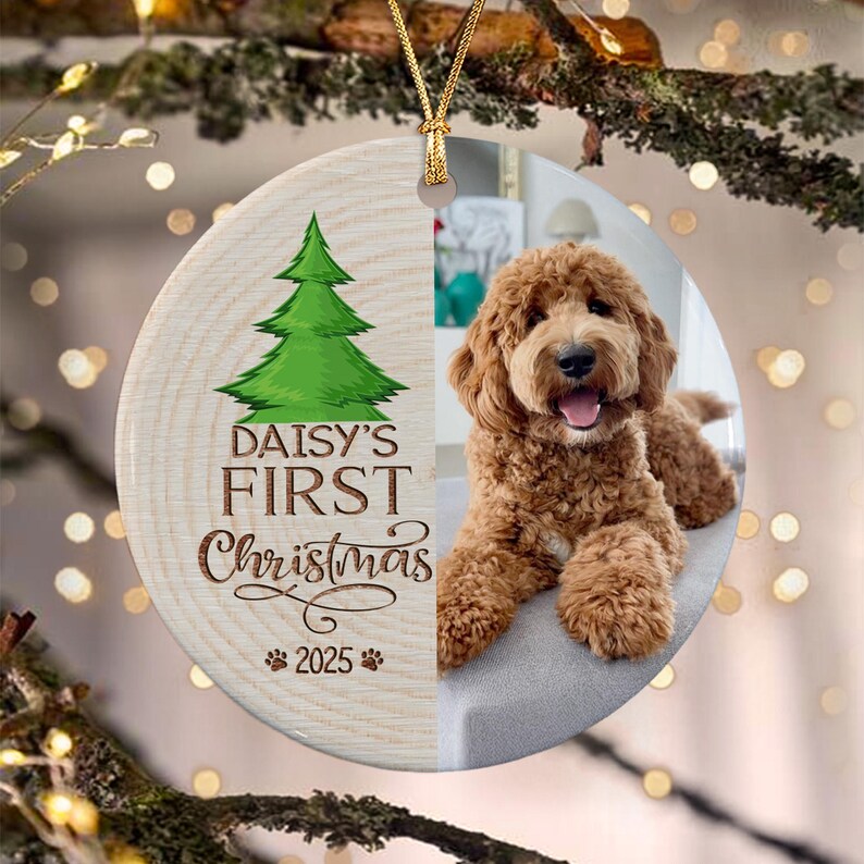 A round ceramic ornament featuring a split design. The left side has a wood grain texture with a green Christmas tree graphic, the text "DAISY'S FIRST Christmas 2025" and two paw prints.