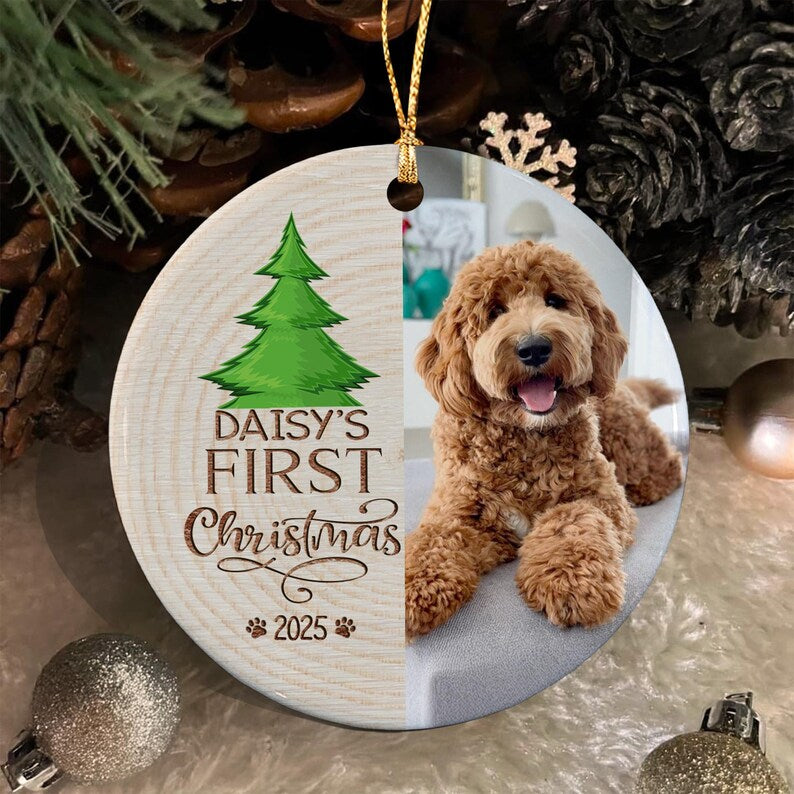 A round ceramic Christmas ornament featuring a split design. The left side has a wood grain texture with a green pine tree illustration and the text "DAISY'S FIRST Christmas 2025".