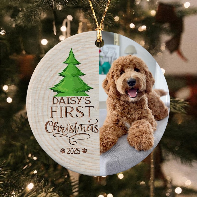 A round ceramic ornament featuring a split design. The left side has a wood grain texture with a green Christmas tree illustration, the text "DAISY'S FIRST Christmas 2025" and two paw prints.