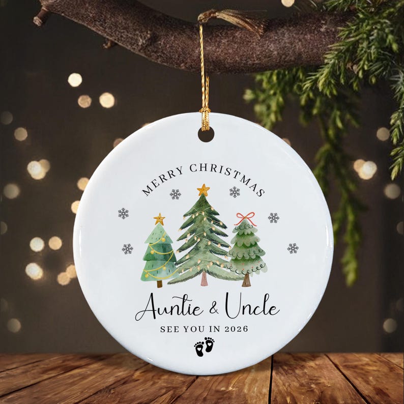 A round white ceramic Christmas ornament featuring a watercolor illustration of three green pine trees with gold stars and snowflakes.