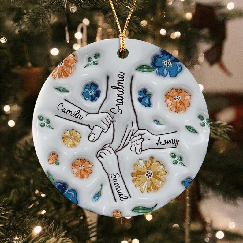 A round white ceramic ornament featuring a central illustration of hands holding together. The text Grandma, Camila, Avery, and Samuel is printed on the surface.