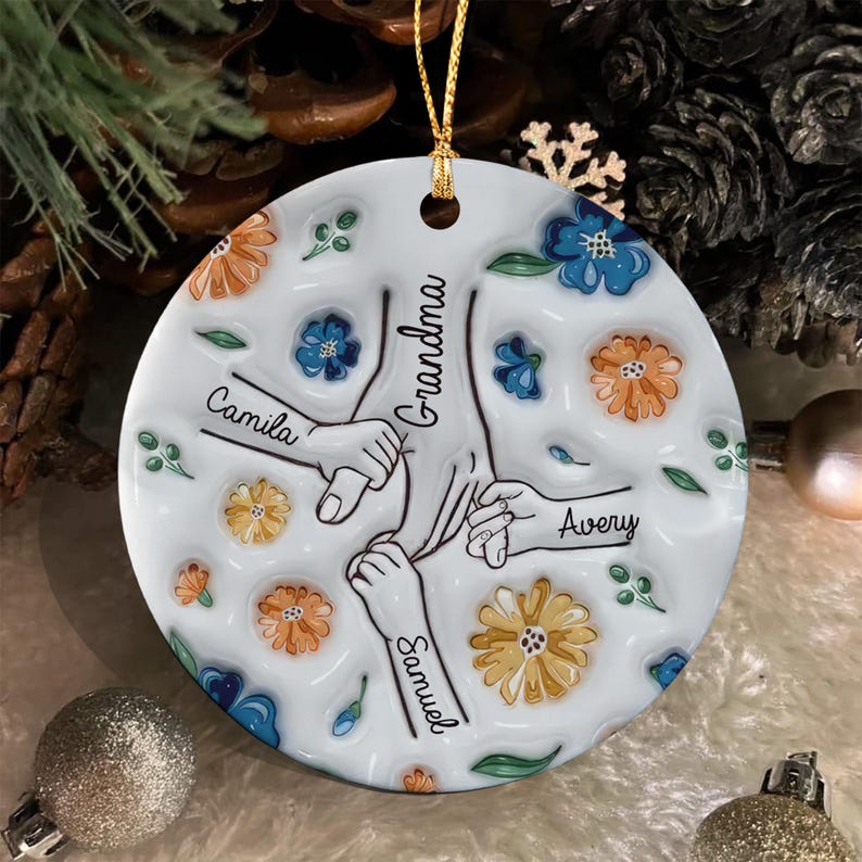 A round white ceramic ornament featuring a central illustration of three hands holding a larger hand. The names Camila, Samuel, and Avery are written near each hand, with Grandma in the center.