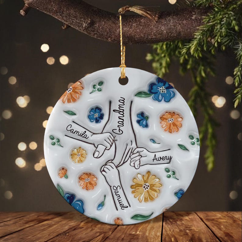 A round white ceramic ornament featuring a central illustration of multiple hands holding onto a central hand labeled Grandma. The names Camila, Avery, and Samuel are written near each hand.